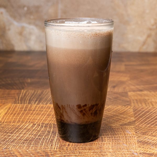 A glass of chocolate milk with foam on top, sitting on a wooden table.