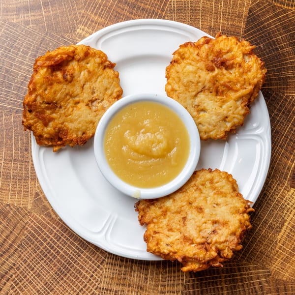 Three golden-brown potato latkes arranged around a small bowl of applesauce on a white plate.