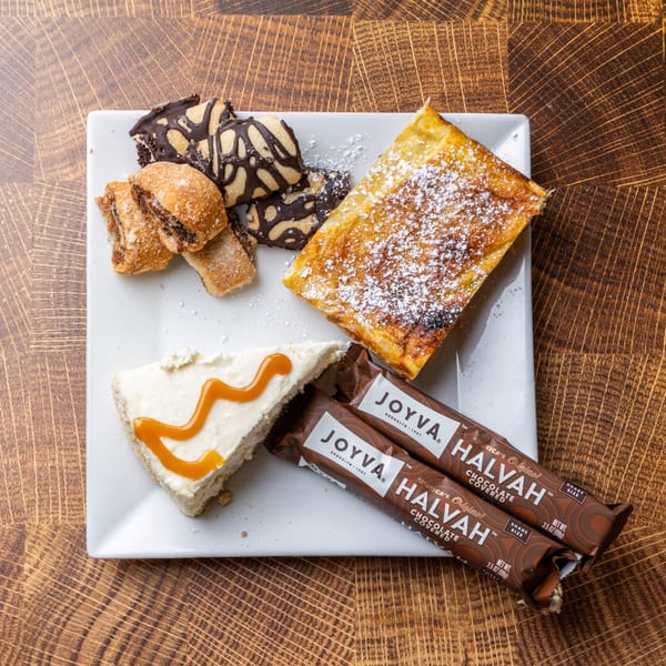 A white plate holds various desserts: cookies, a pastry, cheesecake, and two chocolate-covered halva bars.