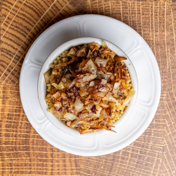 A white bowl of golden couscous topped with caramelized onions, on a wooden table.