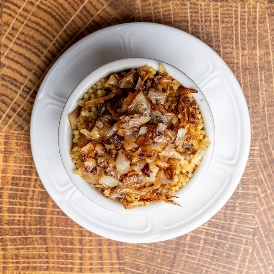 A white bowl of golden couscous topped with caramelized onions, on a wooden table.