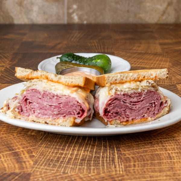 Pastrami Express Reuben sandwich, cut in half, with pickles and sauce on a white plate.