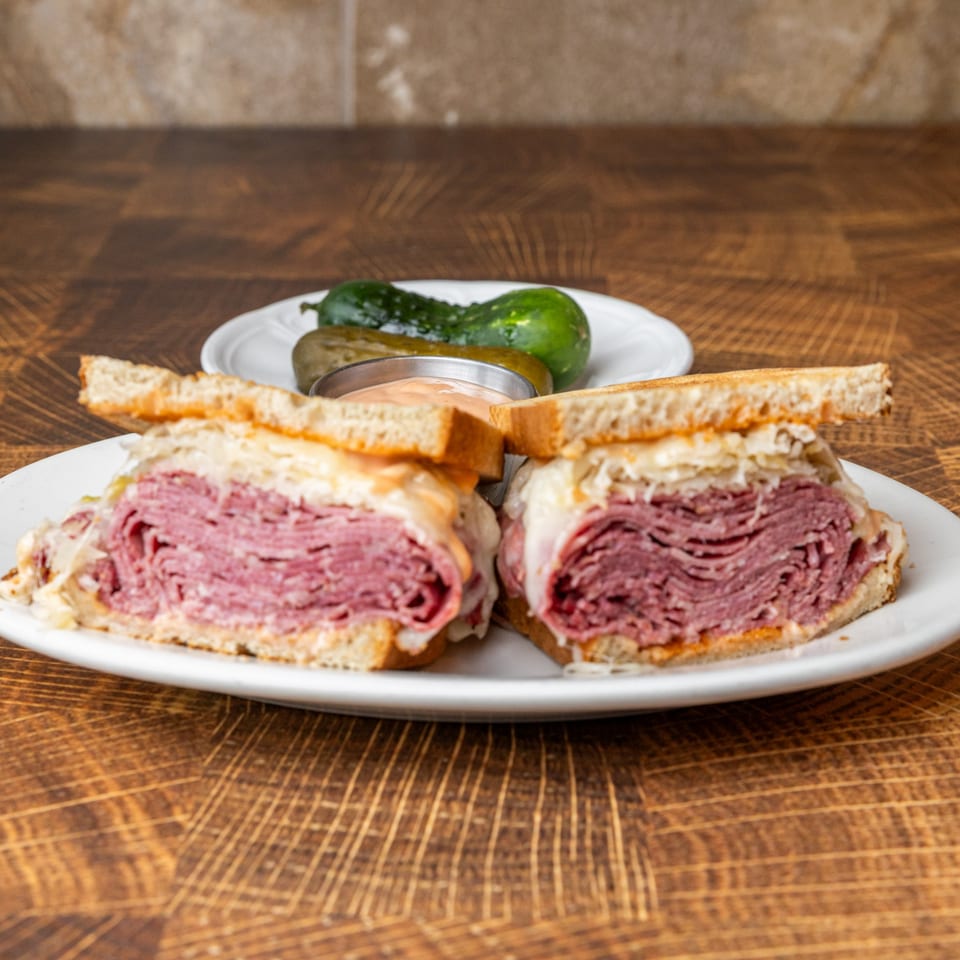Pastrami Express Reuben sandwich, cut in half, with pickles and sauce on a white plate.
