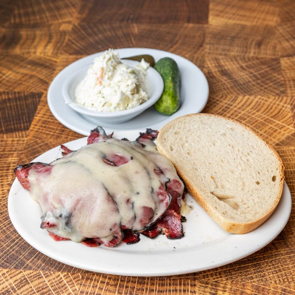 Pastrami Express sandwich with melted cheese, a side of coleslaw, and a pickle on a wooden table.