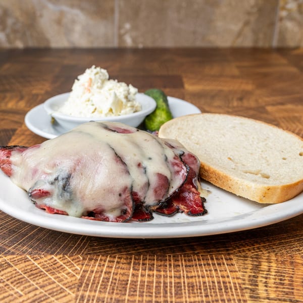 Pastrami Express sandwich with melted cheese, a side of coleslaw, and a pickle on a white plate.