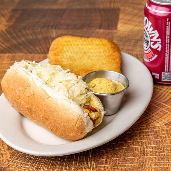 A hot dog with sauerkraut and mustard, a hash brown, and a red soda can on a wooden table.
